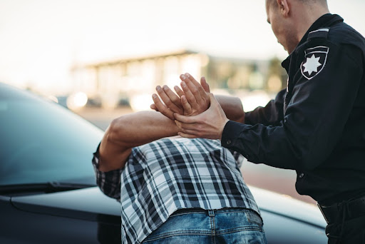 A police officer holds the hands of a man in a plaid shirt behind his head while he bends over a car.